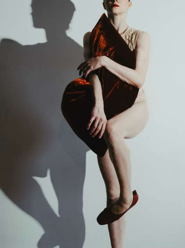 Conceptual studio shot of a slender adult female balancing on one leg while holding a large, plush red velvet pillow against a white wall.