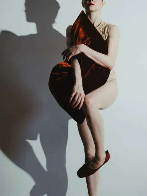 Conceptual studio shot of a slender adult female balancing on one leg while holding a large, plush red velvet pillow against a white wall.