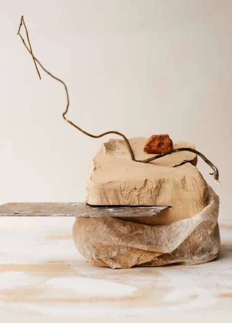 Still life of clay block with metal spatula, small rock, and twig against off-white background creating a minimalistic and artistic composition.