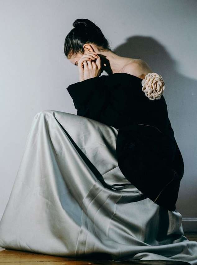 A fair-skinned young woman in a formal black and silvery-gray gown, sits pensively in a photo studio with soft lighting.