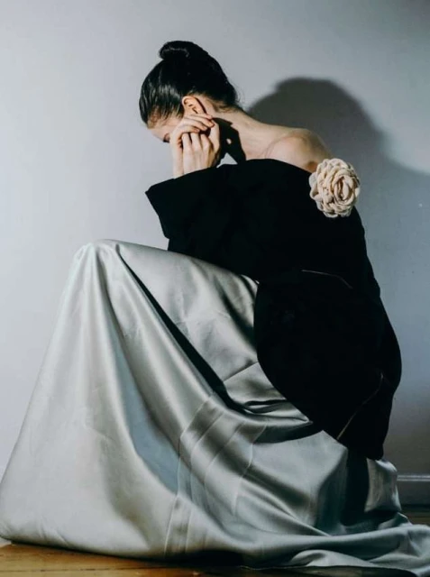 A fair-skinned young woman in a formal black and silvery-gray gown, sits pensively in a photo studio with soft lighting.