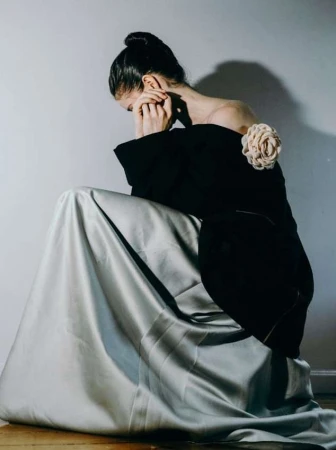 A fair-skinned young woman in a formal black and silvery-gray gown, sits pensively in a photo studio with soft lighting.