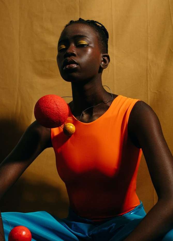 Woman with orange top and blue pants poses with red and yellow spheres on a golden background in a portrait-style studio shot.