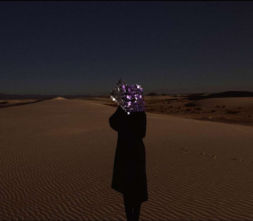 A solitary figure in a long, dark coat stands on a rippled sand dune, head obscured by a reflective cube under a clear dusk sky.