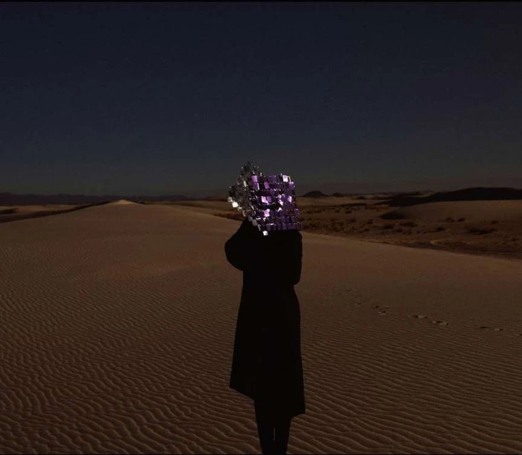 A solitary figure in a long, dark coat stands on a rippled sand dune, head obscured by a reflective cube under a clear dusk sky.