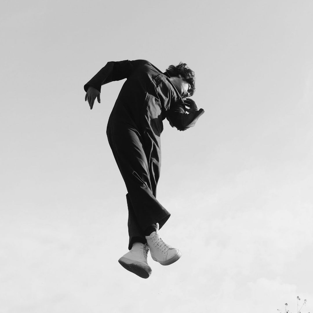 Monochrome portrait of a person jumping in mid-air wearing dark overalls and white high-top sneakers against a light sky