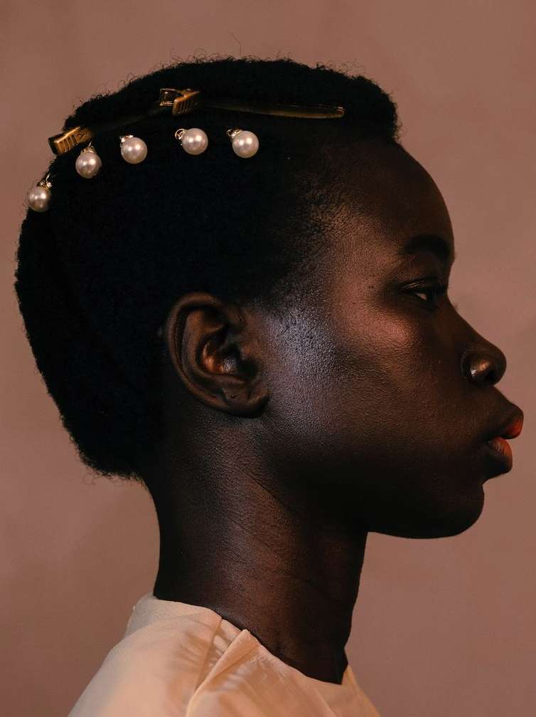 Profile shot of an African American woman with dark skin and short black hair, adorned with pearl-embellished hair clips, against a tan background.