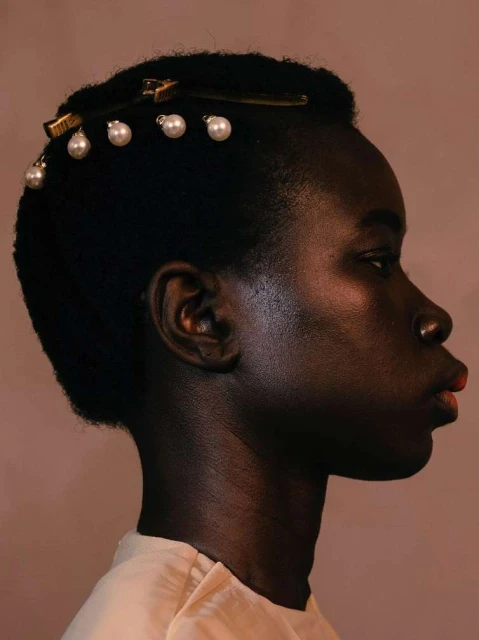 Profile shot of an African American woman with dark skin and short black hair, adorned with pearl-embellished hair clips, against a tan background.