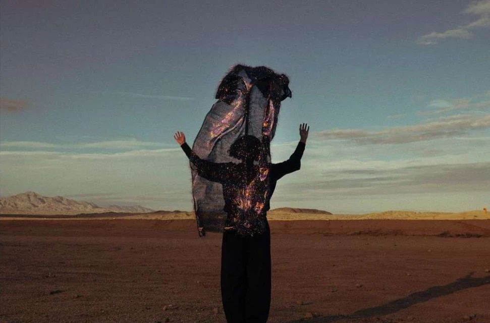 Woman in dark clothing raises arms in desert landscape, wearing glittery jacket and holding reflective fabric against a blue sky.