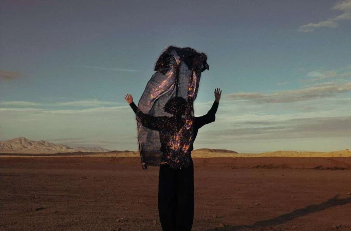 Woman in dark clothing raises arms in desert landscape, wearing glittery jacket and holding reflective fabric against a blue sky.