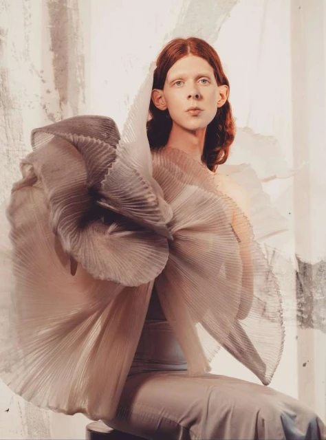 Fashion shot of a red-haired adult in a pale dress with pleated details and a large pleated ruffle against a mottled background.