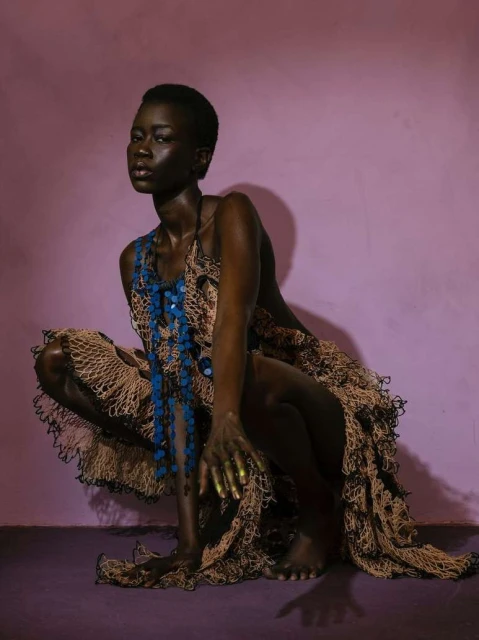 Young Black woman poses in unique dress with blue beaded necklace against a textured purple background in an eye-level medium shot.