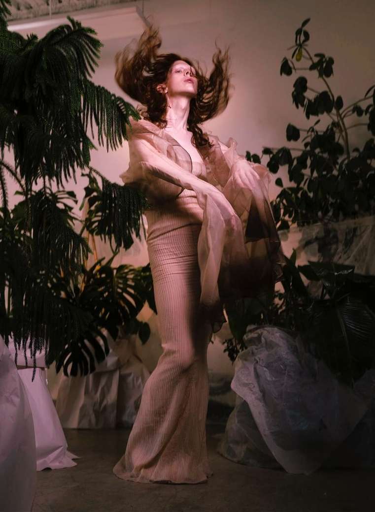 Full shot of woman posing in textured maxi dress and sheer robe, surrounded by green plants in a studio with soft lighting.