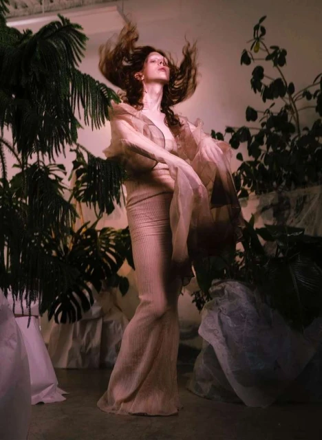Full shot of woman posing in textured maxi dress and sheer robe, surrounded by green plants in a studio with soft lighting.