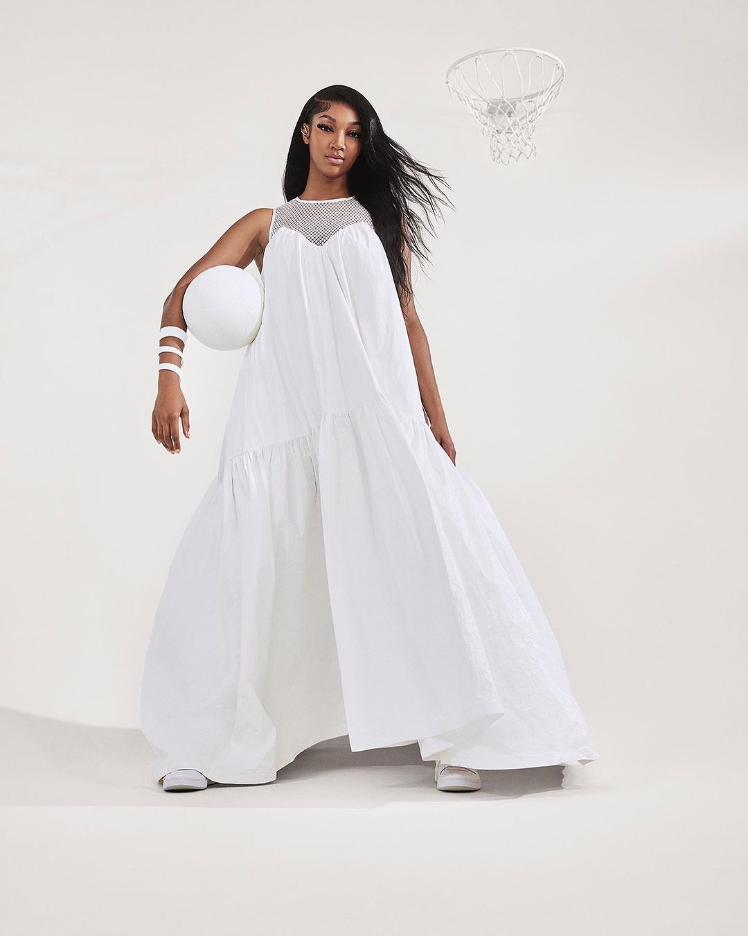 A stylish African American woman in a white dress poses with a basketball in a bright photo studio with a basketball hoop in the background.