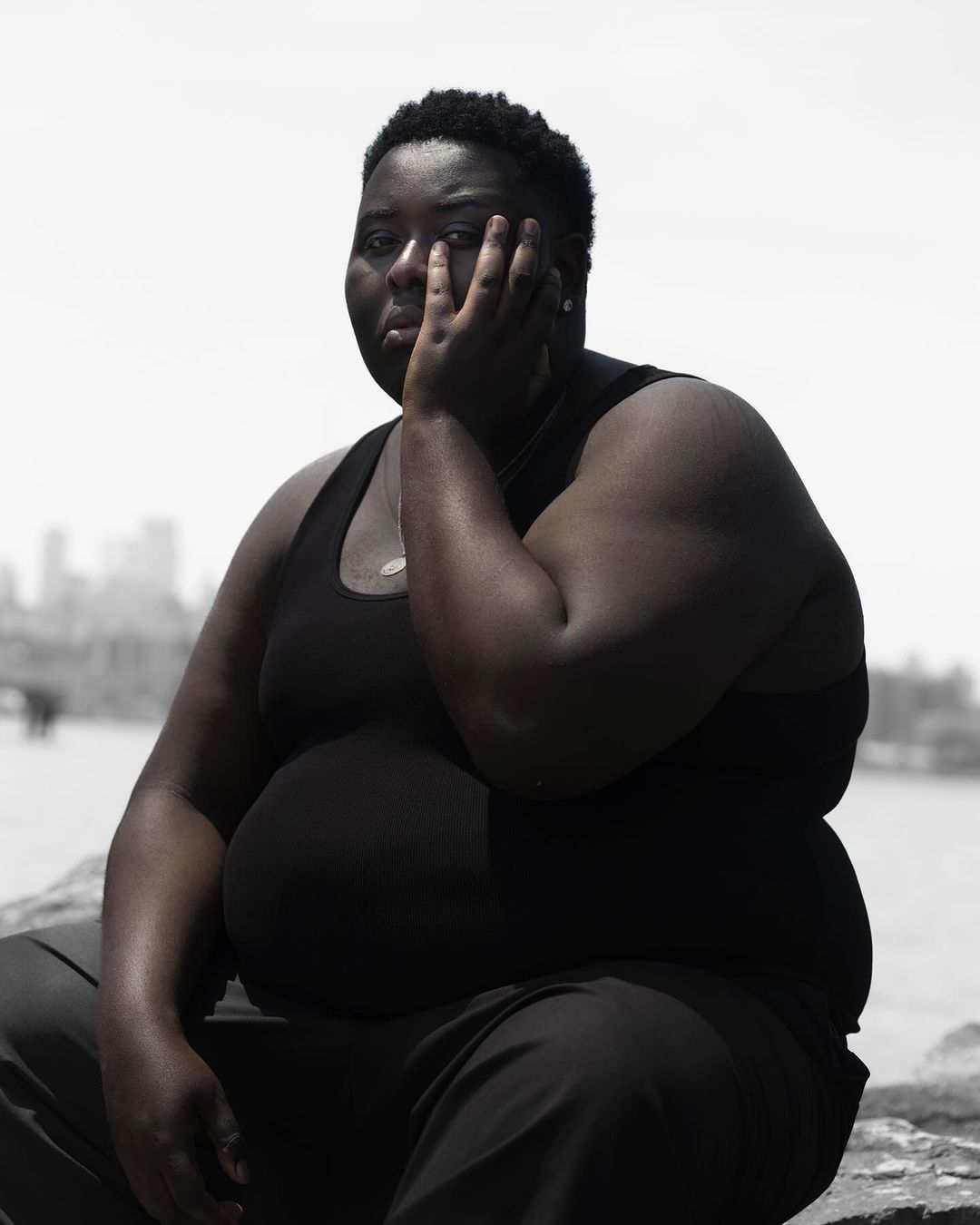 Monochromatic portrait of an African American adult female with short hair, hand to face, posing outdoors near a city skyline.