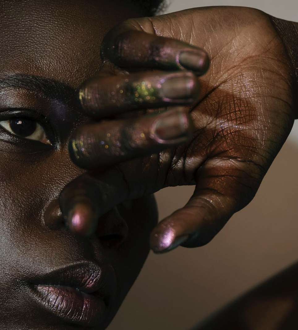 Close-up of a woman's face, her hand partially covering it, showcasing purple-toned skin and makeup against a soft, neutral background.