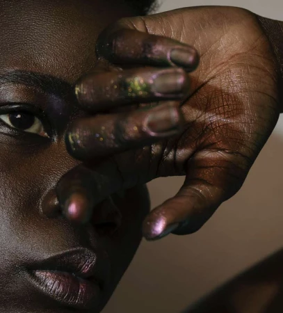 Close-up of a woman's face, her hand partially covering it, showcasing purple-toned skin and makeup against a soft, neutral background.