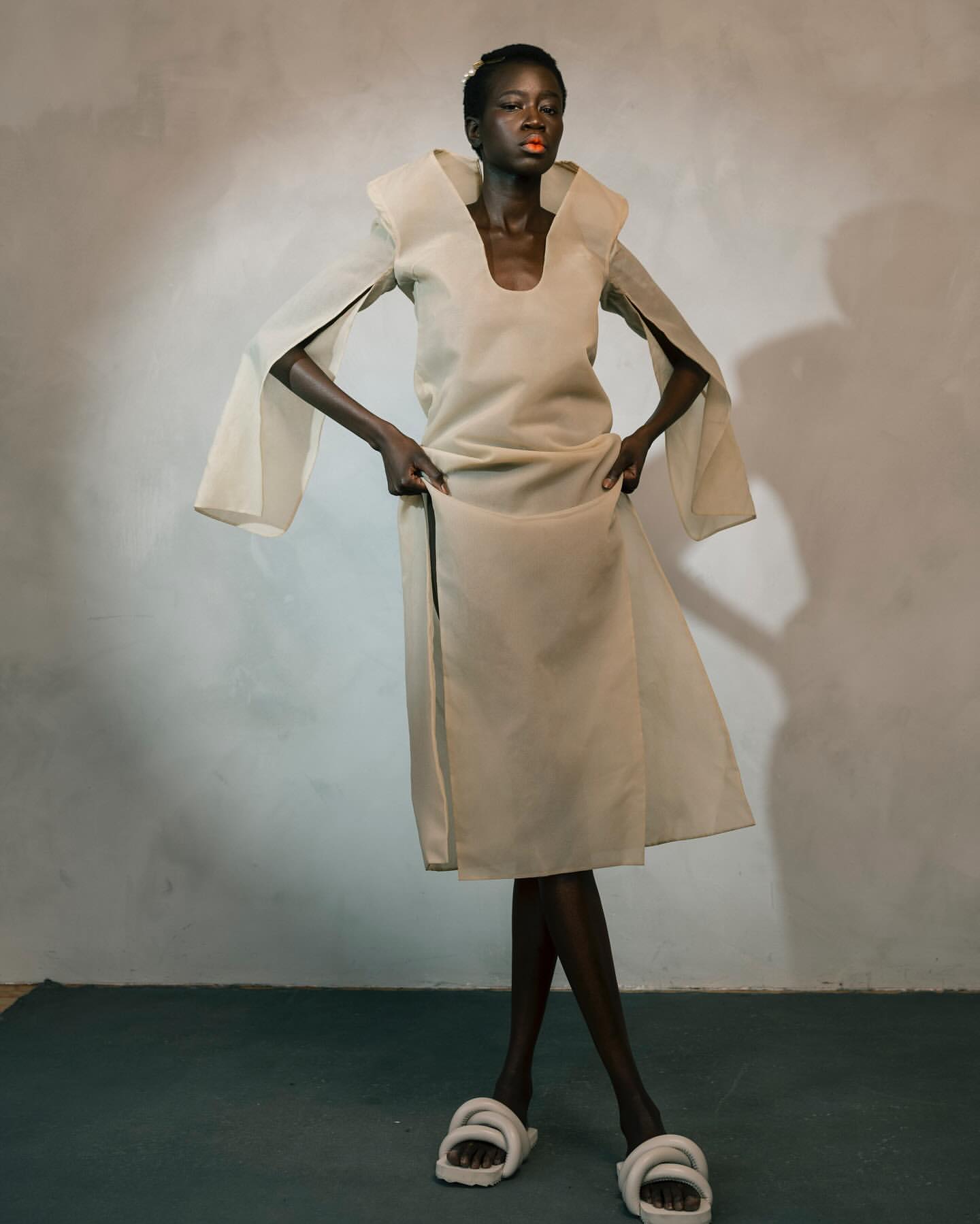 Fashion model in a beige dress with wide sleeves poses in a studio with textured white walls and large white slide sandals.