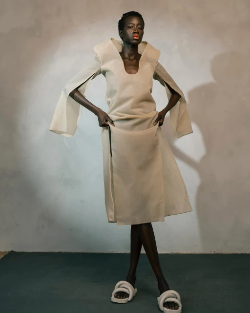 Fashion model in a beige dress with wide sleeves poses in a studio with textured white walls and large white slide sandals.