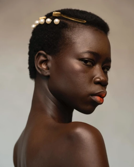 Portrait of a dark-skinned woman with pearl-accented hair clips, striking red-orange lipstick, and a neutral background in a beauty editorial.