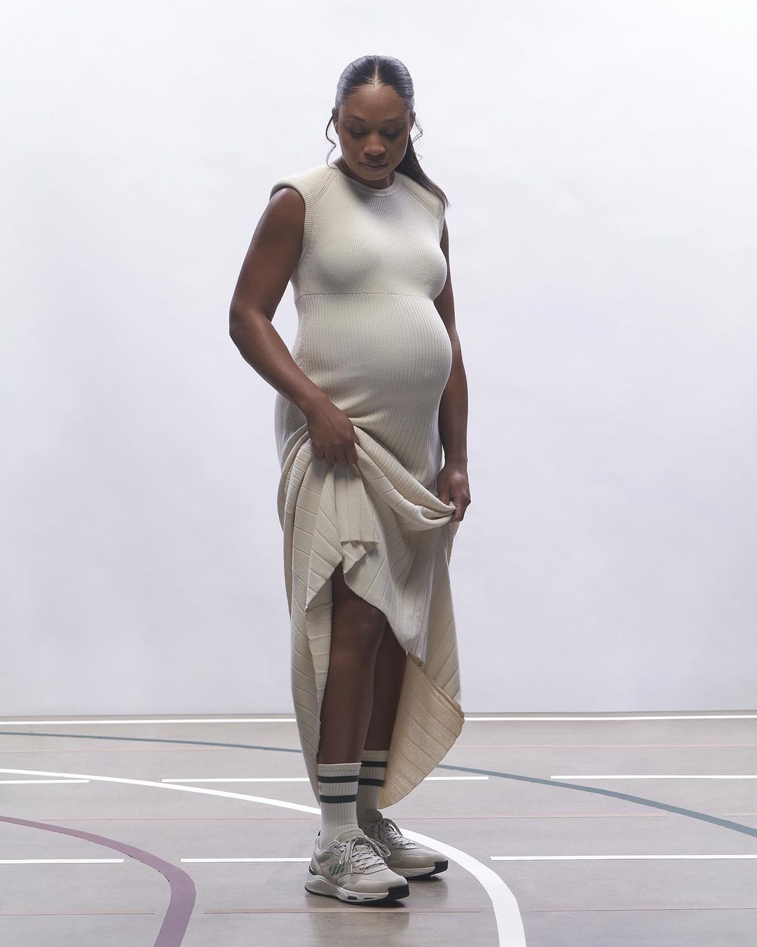 Pregnant African-American woman in a cream-colored dress standing in a gymnasium with athletic shoes, facing downward, looking at her belly.