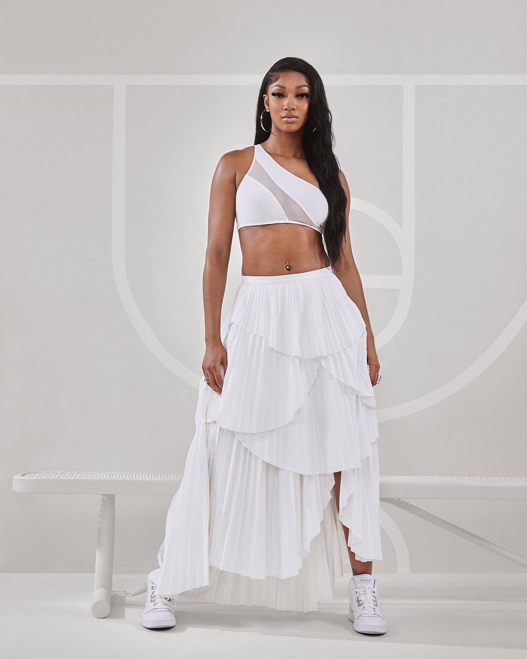 A fashionable woman stands in a white one-shoulder crop top and tiered skirt with sneakers, posing in front of a minimal indoor backdrop.