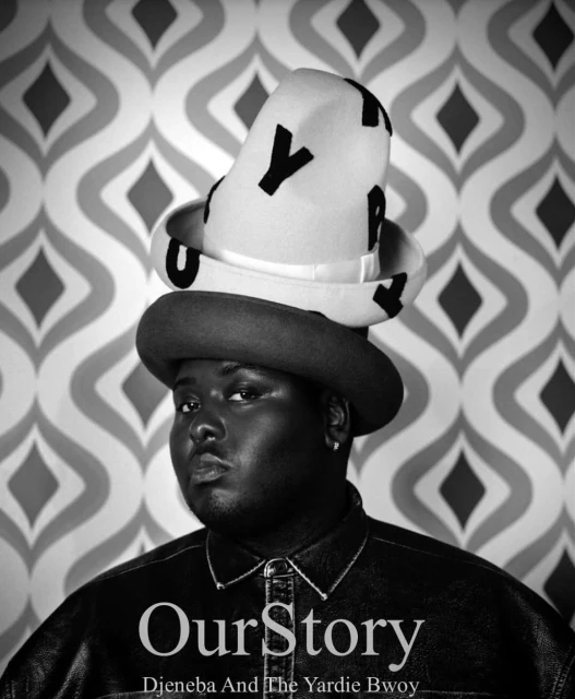 Studio portrait of a person with dark skin wearing a dark jacket and a layered hat against a patterned gray and white wallpaper.