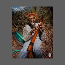 Black man in striped overalls holding a wooden instrument outdoors with spiky foliage background