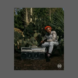 Man in orange beanie and patterned overalls sitting outdoors beside a large boombox with a dense cactus garden background.