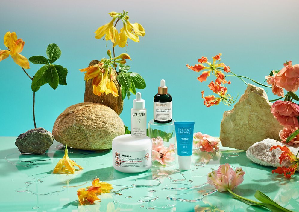 Still life featuring an arrangement of skincare products from Biossance, Caudalie, and Perricone MD with rocks and colorful flowers on a blue surface.