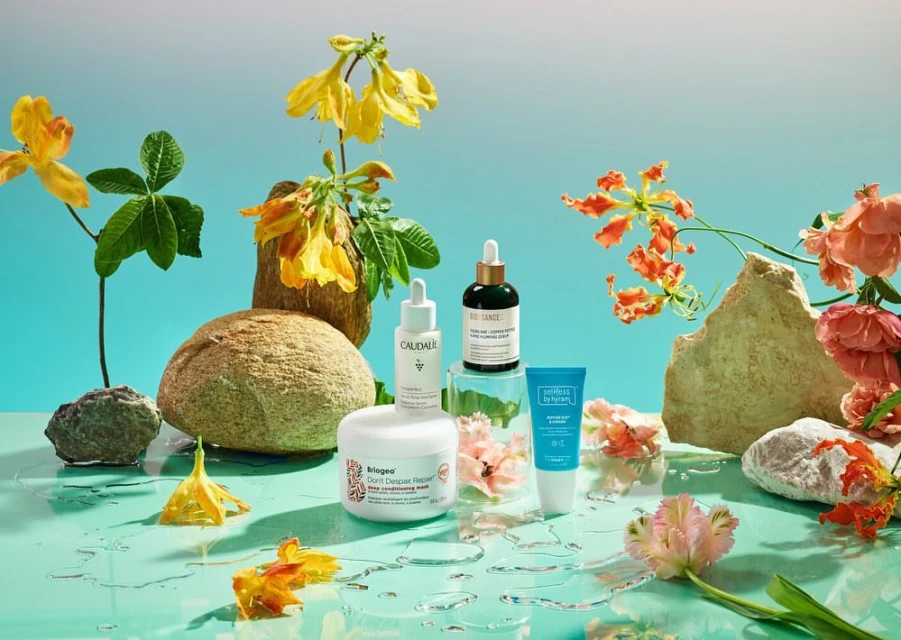 Still life featuring an arrangement of skincare products from Biossance, Caudalie, and Perricone MD with rocks and colorful flowers on a blue surface.