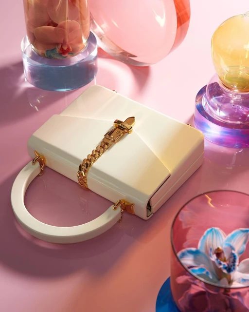 Elevated studio shot of a white handbag with a gold chain and round handle, styled with glass vases on a pink surface.