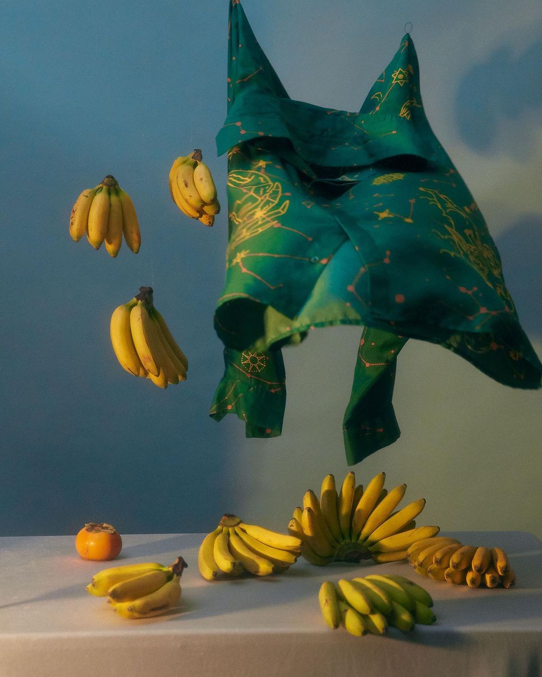 Still life food photography featuring bunches of bananas, a persimmon fruit, and a patterned green cloth set on a white surface.
