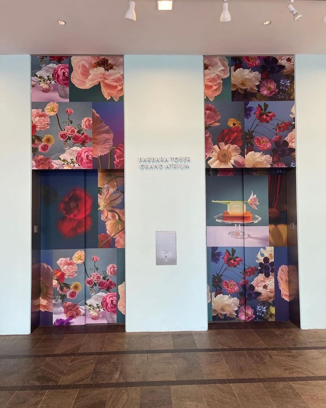Elevator doors with floral print artwork in an atrium with wooden floors and light walls under ceiling lights. Contemporary floral design.