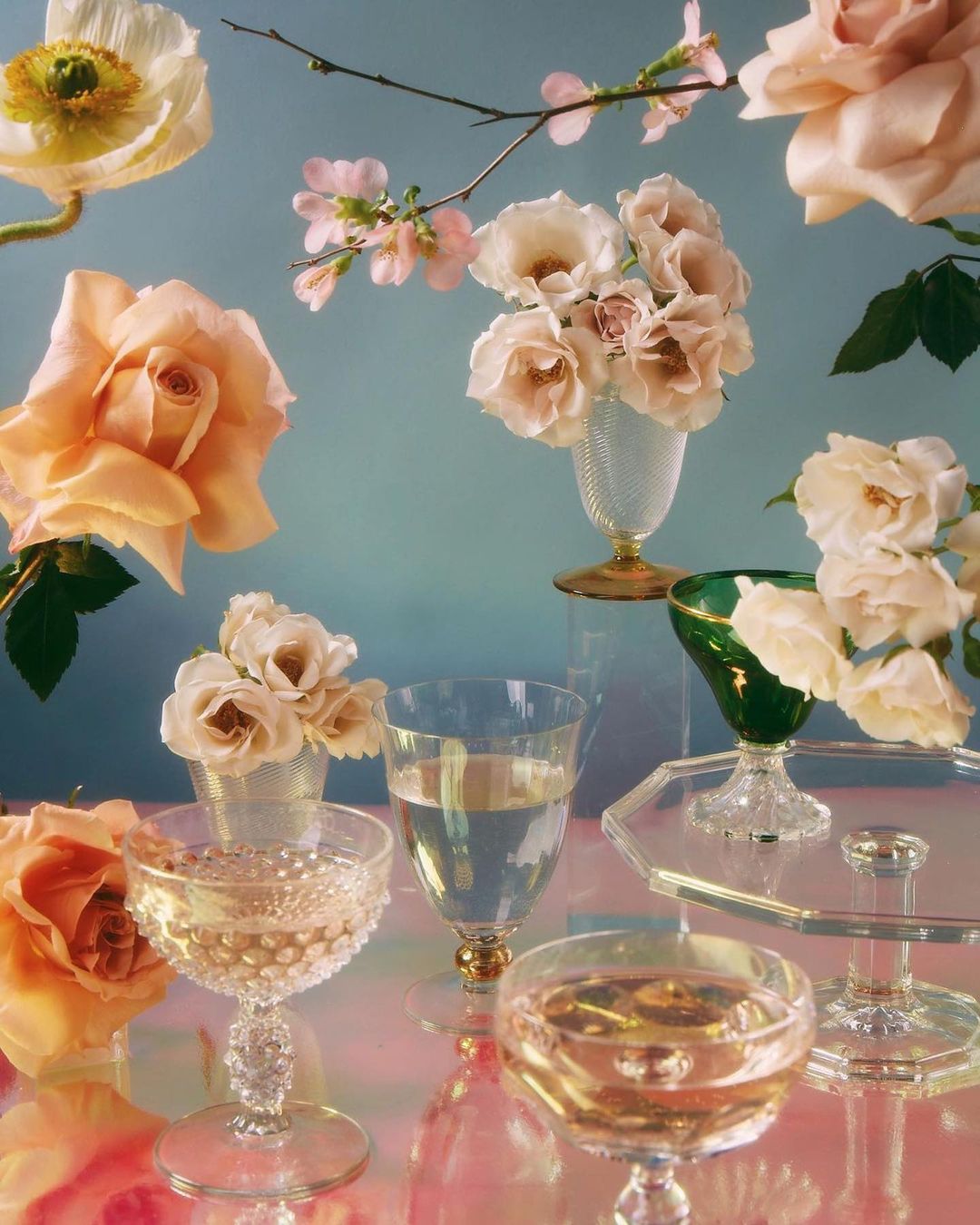 Floral still life featuring champagne flutes, glass vases, and pastel roses against a light blue backdrop. Elegant arrangement.