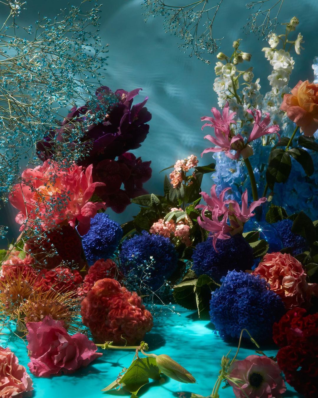Floral arrangement featuring a variety of colorful flowers, including roses, peonies, and lilies, set against a textured teal background.