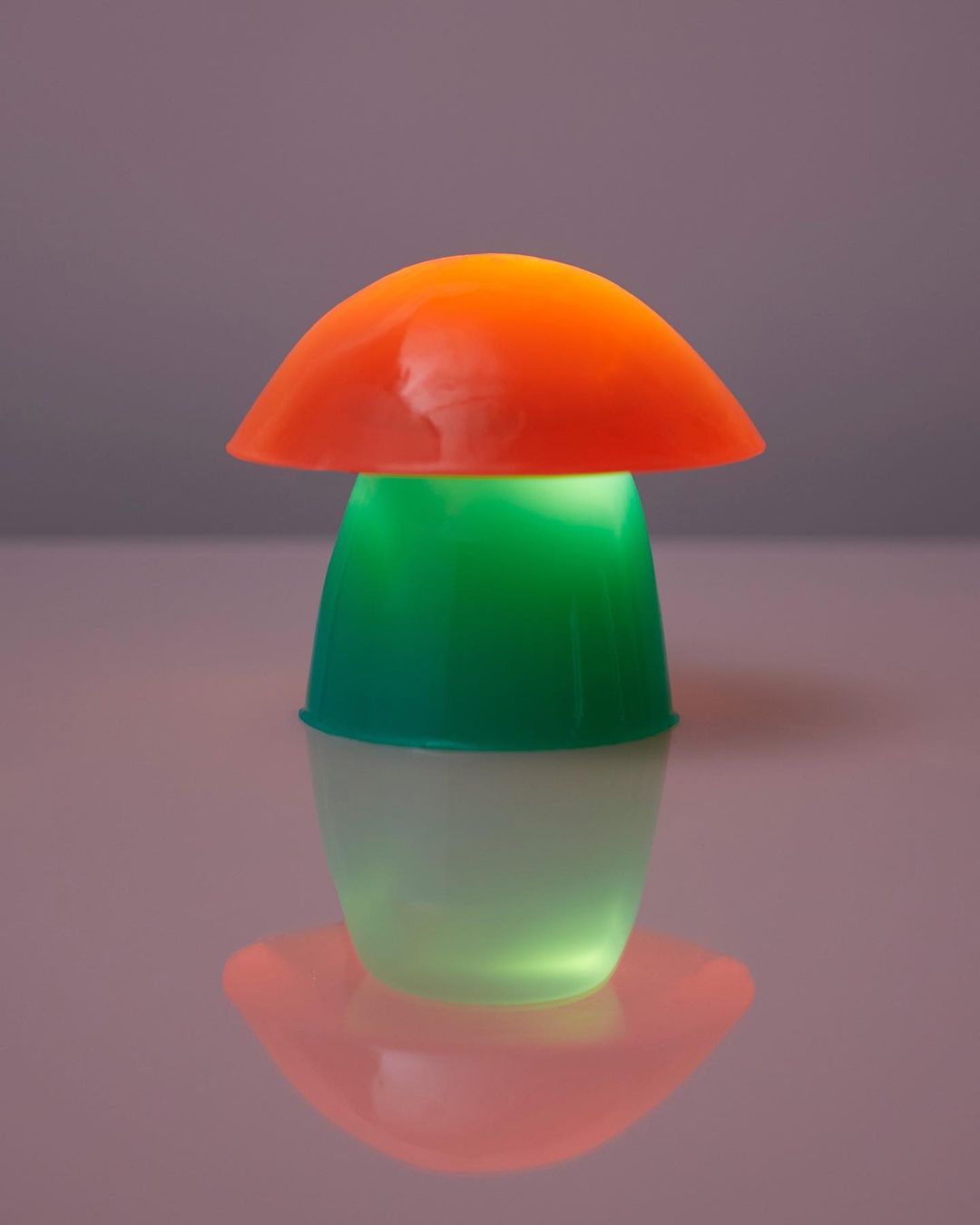 A mushroom-shaped lamp shines with orange, green, and white light, creating a colorful and playful illumination on a reflective surface.