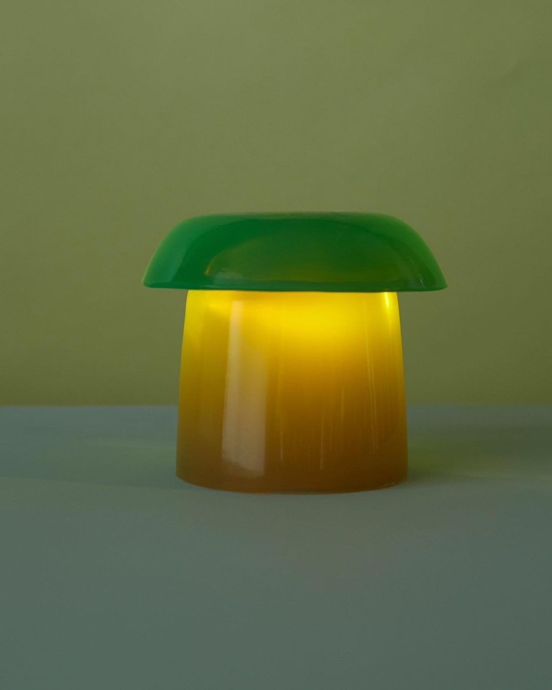 Illuminated green and yellow mushroom lamp casting a warm glow, set against a muted yellow background. Table lamp emitting light.