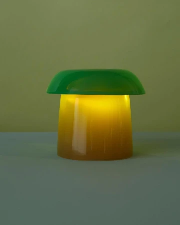 Illuminated green and yellow mushroom lamp casting a warm glow, set against a muted yellow background. Table lamp emitting light.