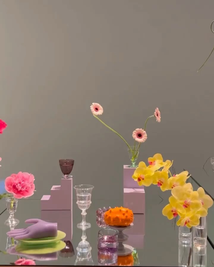 Elegant still life arrangement with pink and yellow flowers, glassware, and decorative objects on a reflective surface against a grey wall.