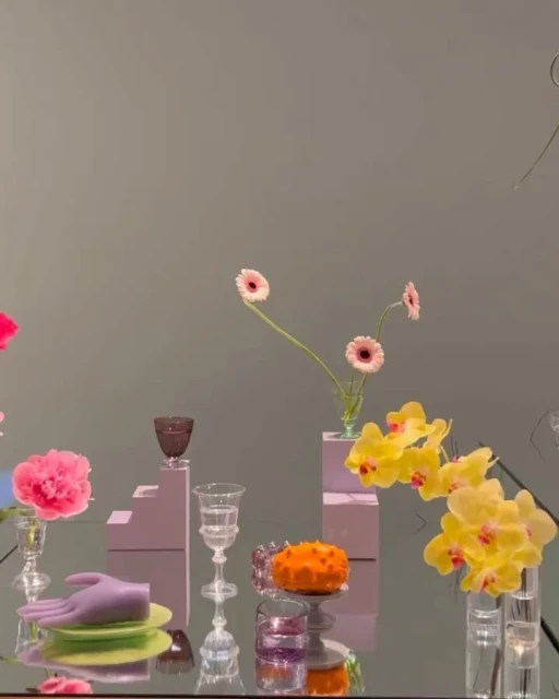 Elegant still life arrangement with pink and yellow flowers, glassware, and decorative objects on a reflective surface against a grey wall.