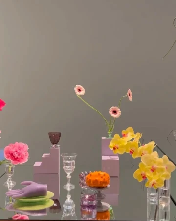 Elegant still life arrangement with pink and yellow flowers, glassware, and decorative objects on a reflective surface against a grey wall.