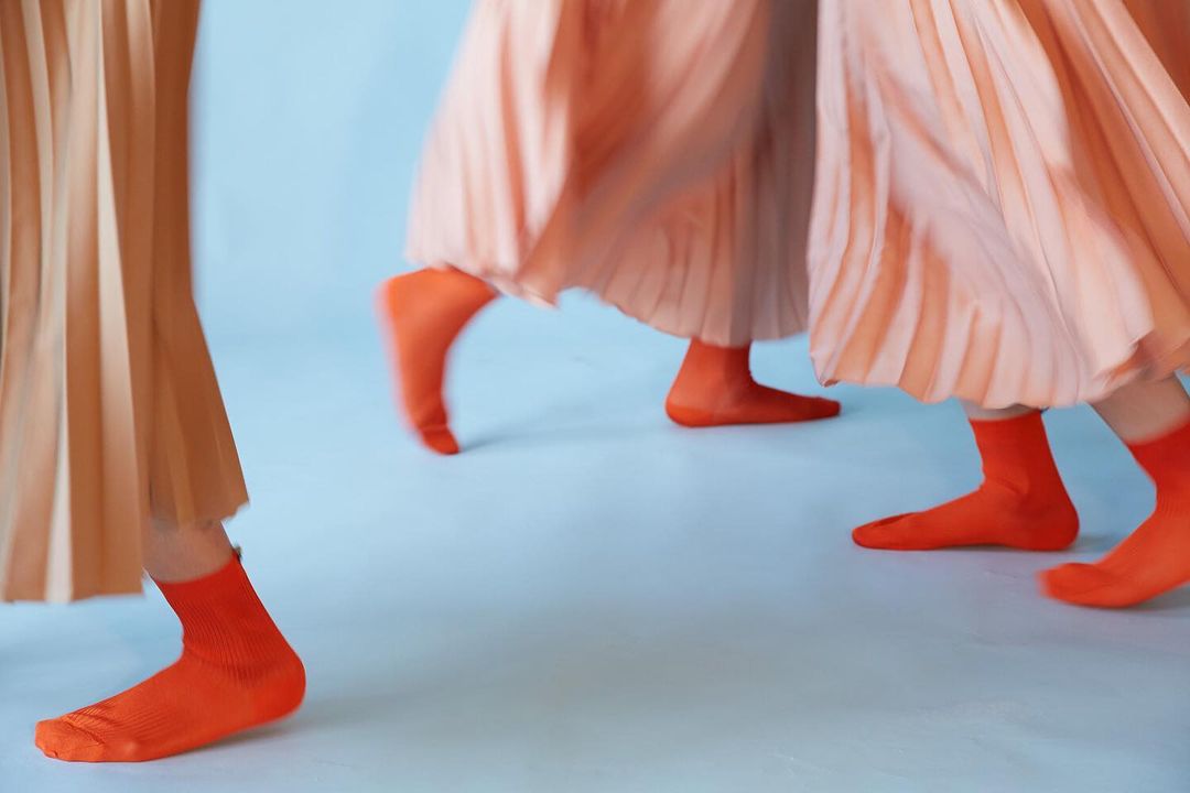 Three people in pleated skirts and orange socks walk against a light blue background, focusing on legs and feet in a fashion setting.