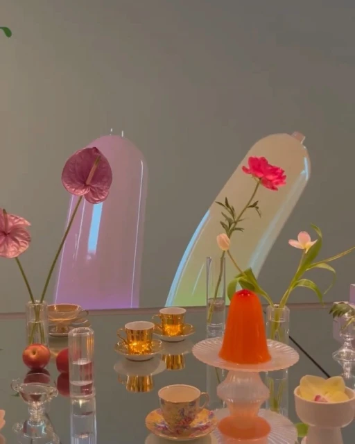 Still life tabletop scene with pink Anthurium, red peony in translucent containers, assorted cups, decorative objects on reflective surface.