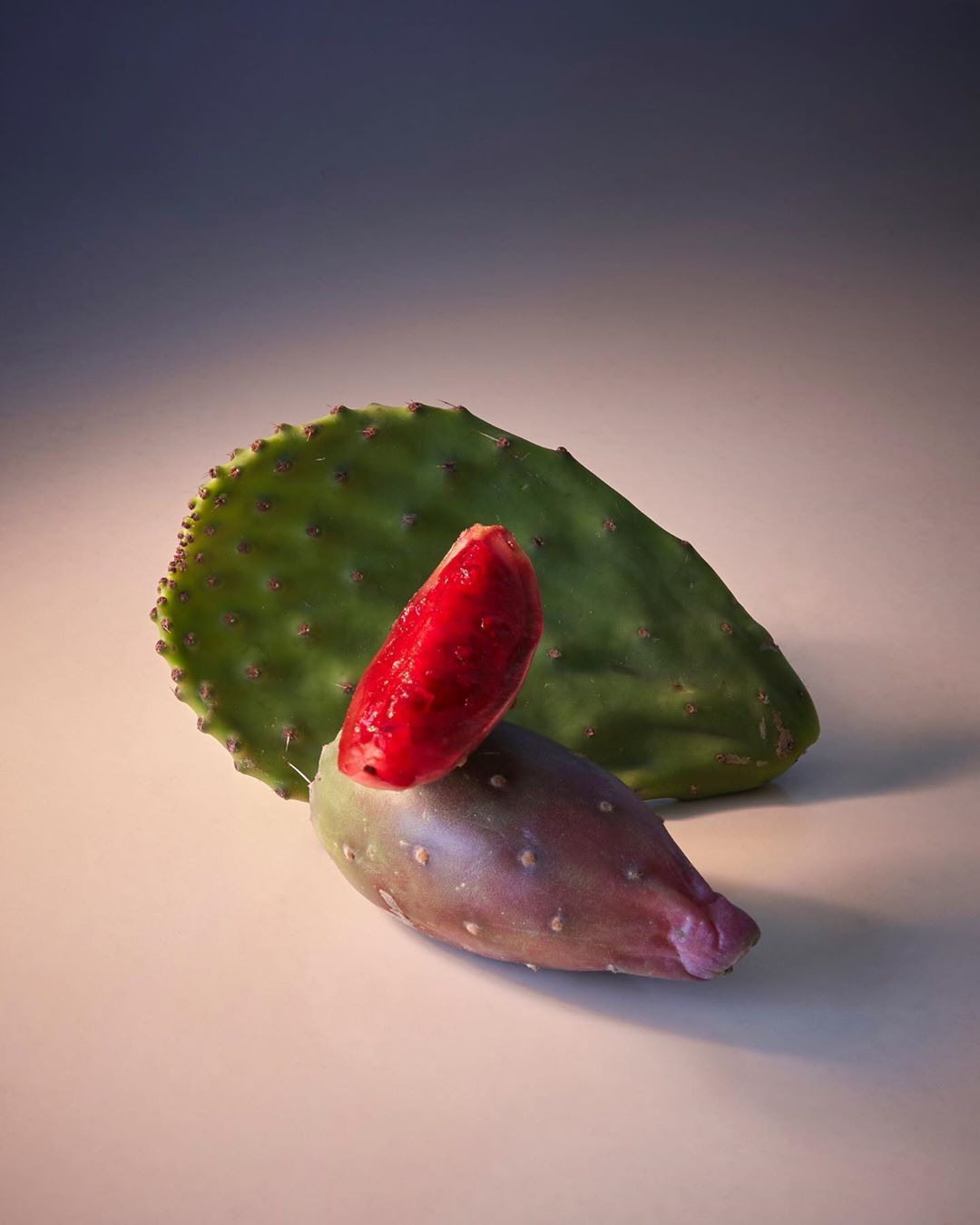 Still life of green prickly pear cactus pad with ripe red fruit segment and purple cactus fruit on a peach-colored surface.