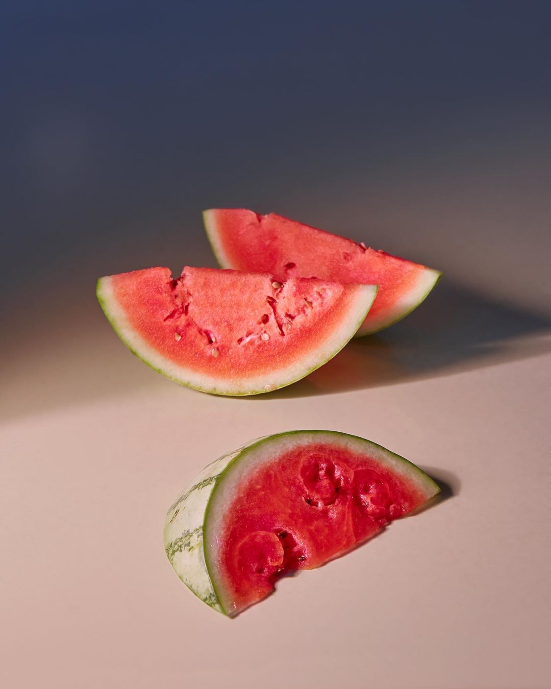 Three watermelon wedges with red flesh and green rind are arranged on a light surface against a gradient blue backdrop, casting shadows.