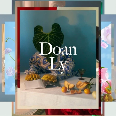 Abstract still life shot of a large green leaf positioned over a person's head, with fruit and book on a draped white table.