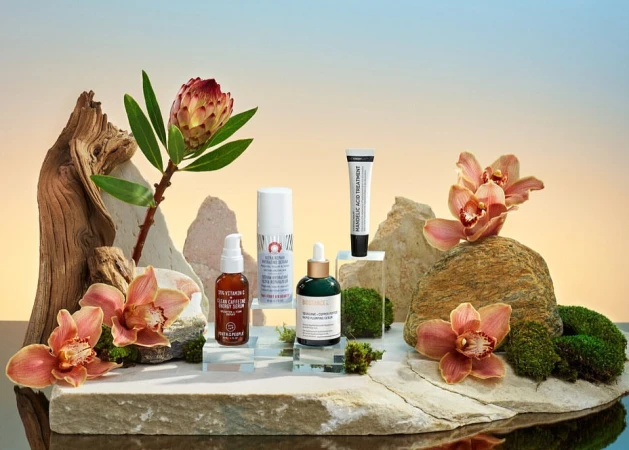 Still life of skincare products arranged with flowers, stones, and wood against a gradient background in a beauty product display.