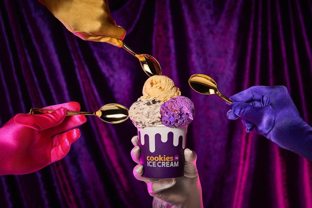 Ice cream in a cup with four gloved hands holding golden spoons, set against a draped purple background in a studio.